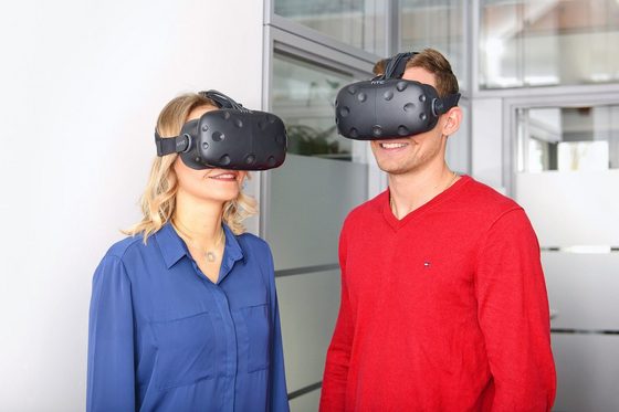 Ein Mann und eine Frau mit aufgesetzten Virtual-Reality-Brillen stehen in einem Raum und lächeln.